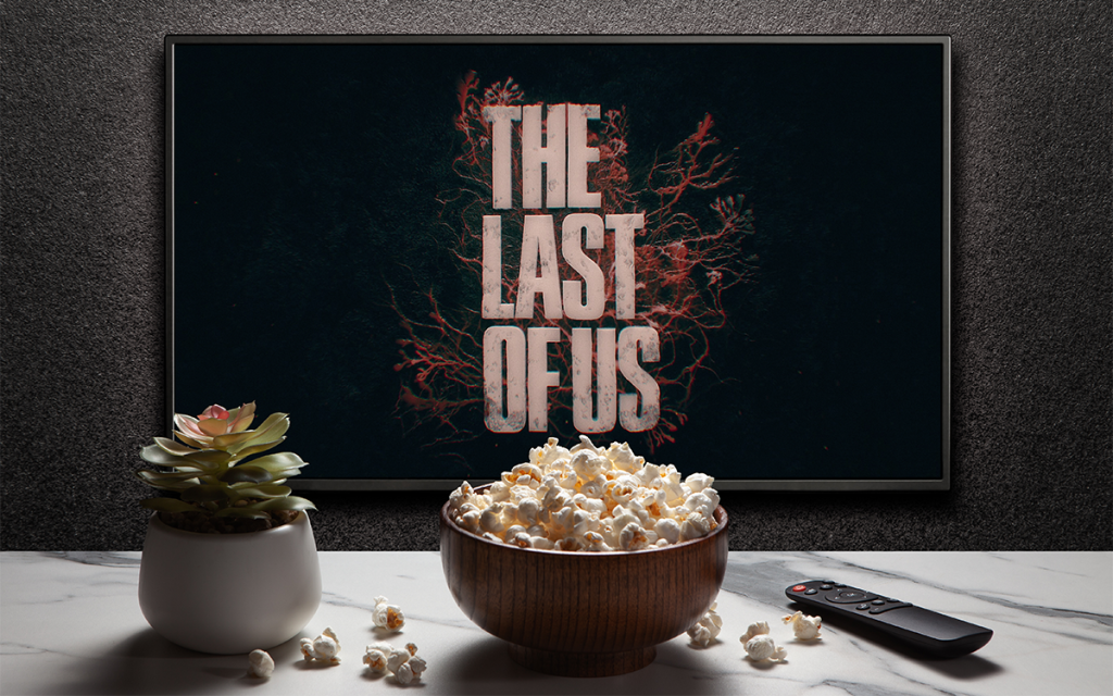 televisão com logo de introdução da série de The Last of Us, uma das séries de maior sucesso da Max, com balde de pipoca, controle remotro e vaso de planta em cima de uma mesa