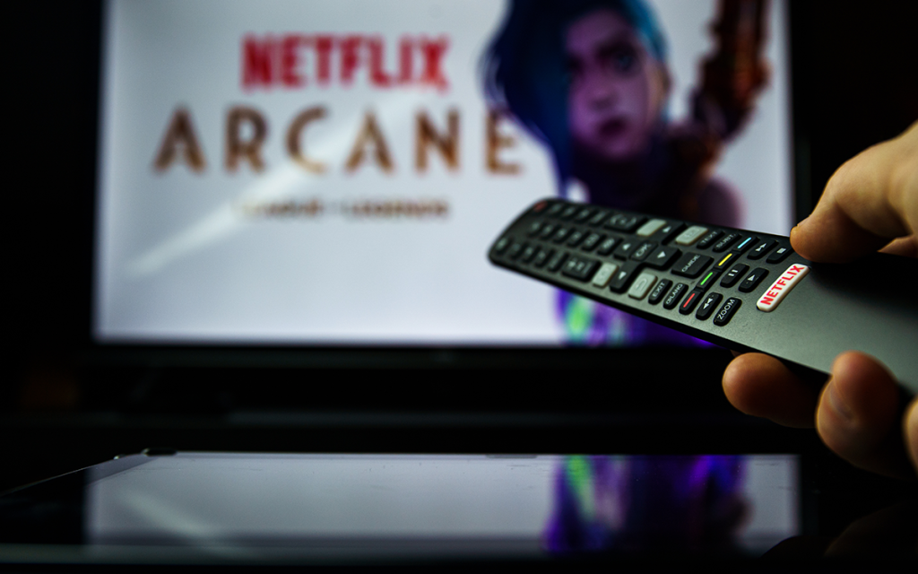 Mão com controle remoto apontando para televisor com a capa de Arcane, uma das séries de jogos mais populares já lançadas.