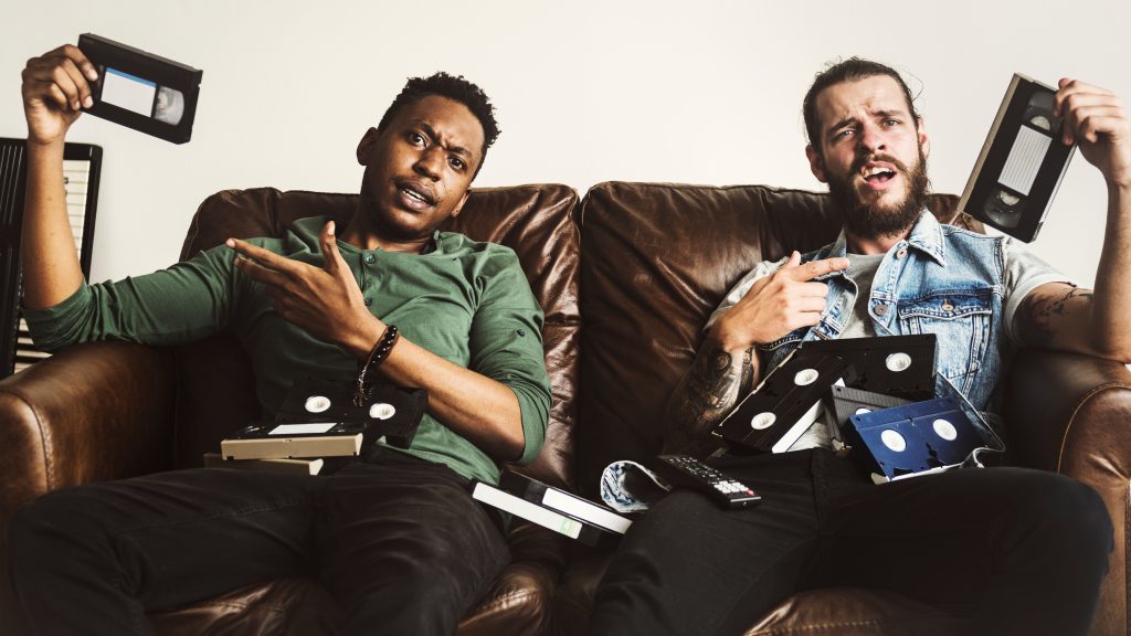 dois homens sentados no sofá com várias fitas cassete representando um dos principais hábitos de consumo dos gamers que são os filmes e séries