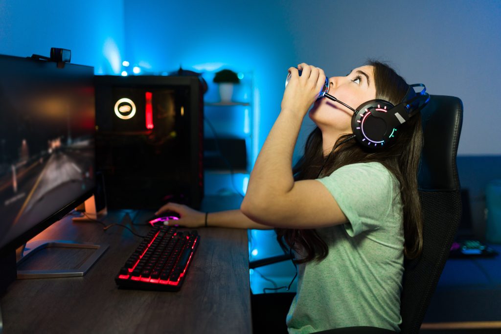 mulher jogando video-game no pc com headset bebendo uma latinha de bebida representando os hábitos de consumo dos gamers em relação a bebidas