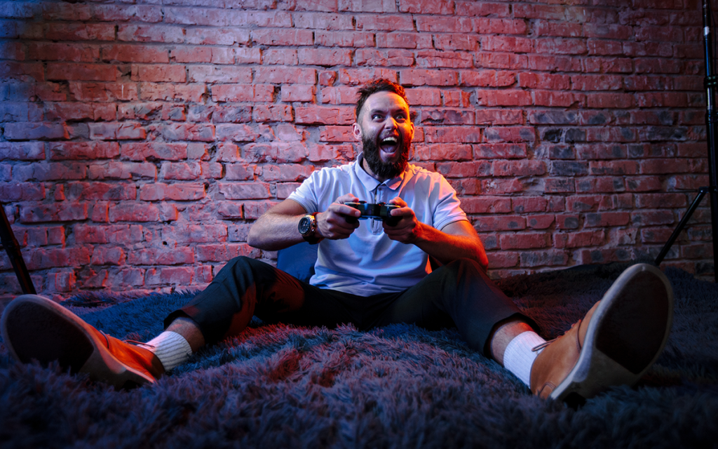 Homem gamer sentado no chão em tapete com expressão de empolgação e ao fundo uma parede de tijolos