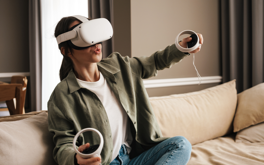 mulher sentada em sofá de casa jogando vídeo-game usando dois controles e um óculos de realidade virtual