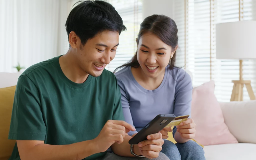 Dois jovens asiáticos sorrindo e olhando para um telefone celular juntos. Eles estão sentados em um sofá em uma sala bem iluminada, o homem vestindo uma camiseta verde e a mulher uma camiseta lilás. Eles parecem estar compartilhando um momento agradável.