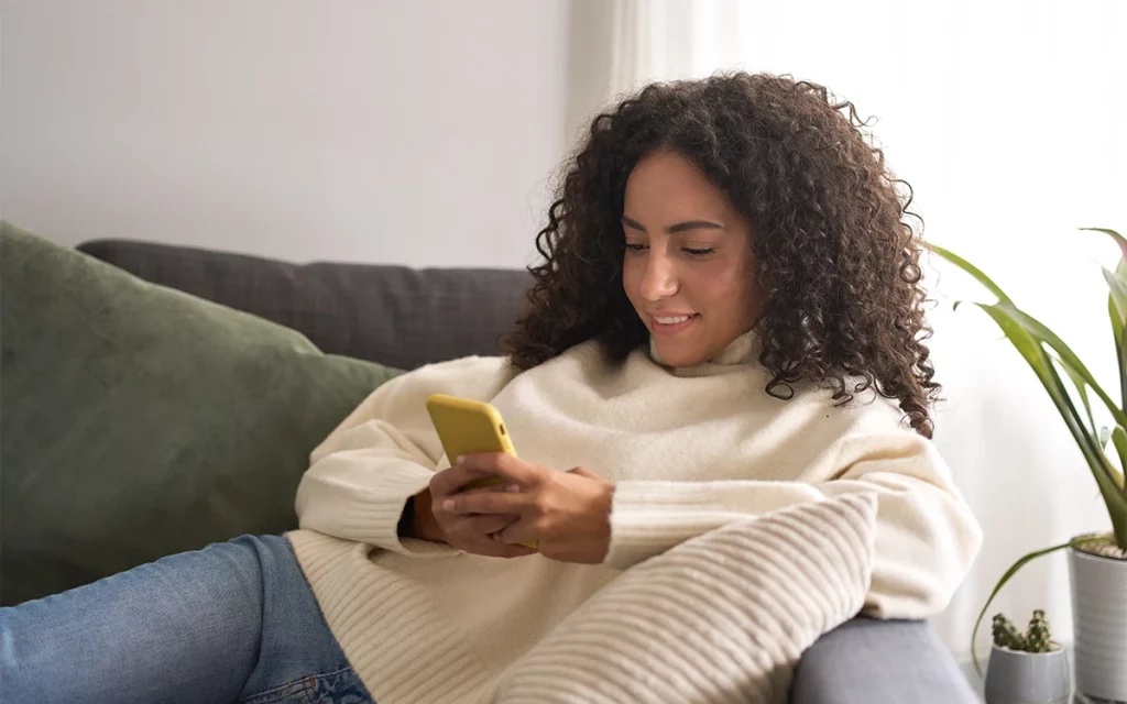 Uma mulher com cabelo encaracolado e vestindo um suéter branco aconchegante está concentrada olhando para seu telefone celular. Ela está sentada confortavelmente em uma sala de estar com um sofá e plantas verdes ao redor, criando uma atmosfera caseira.