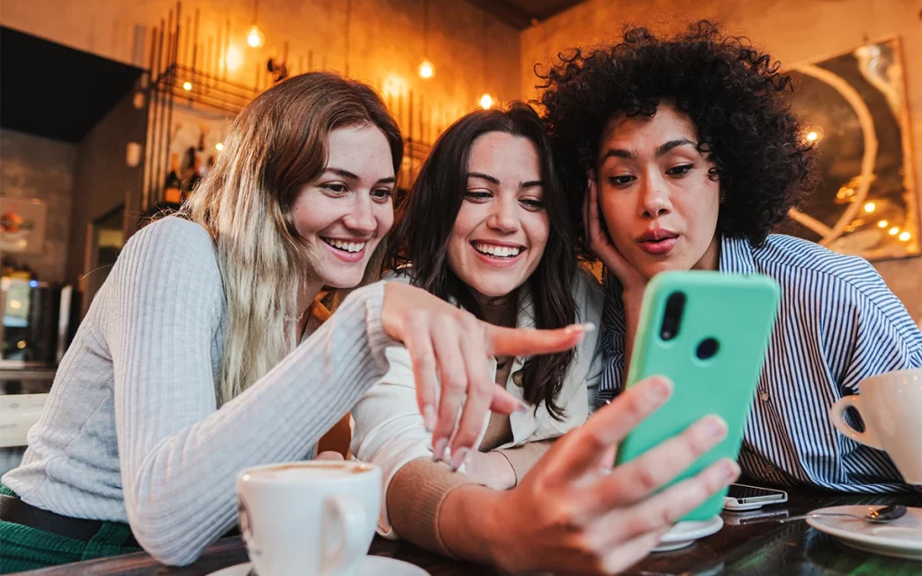 Três amigas compartilhando um momento de alegria olhando para um smartphone, possivelmente experimentando aplicativos juntas através de um aplicativo móvel.
